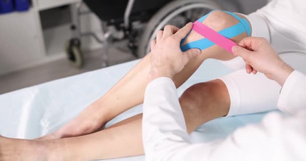 Doctor lijmen kinesiotape op patiënt zere knie in kliniek closeup 4k film slow motion - Video