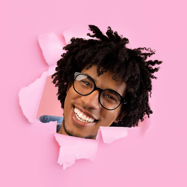 Funny cheerful millennial black man in glasses looks through hole in pink paper, square - Foto, Imagen