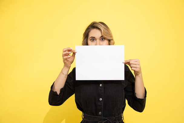 Egy fiatal nő, aki fehér, üres papírlap mögé bújik a felirathoz. Stúdiófelvétel sárga háttérrel. Felismerhetetlen személy. - Fotó, kép