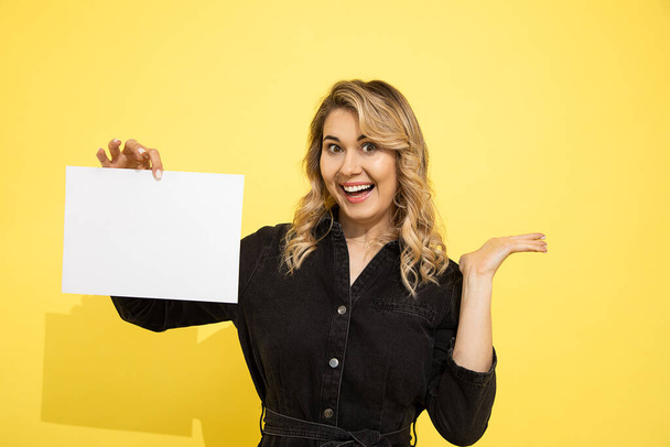 young beautiful blonde woman in black dress holds empty sheet for inscription on yellow background in hands pointing with hand, smiles and laughs happily on yellow background in studio - Фото, изображение