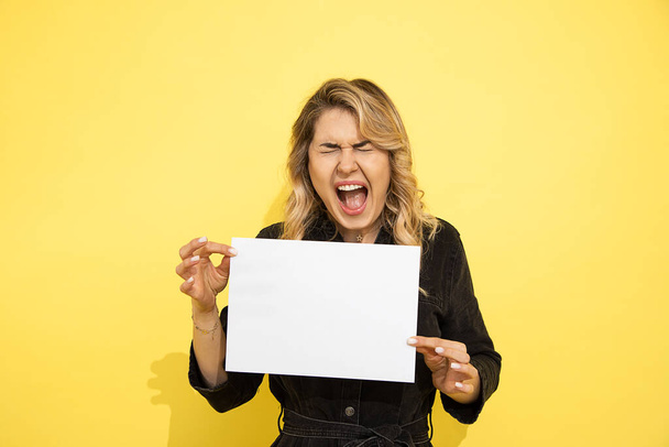  Портрет молодой красивой женщины громко кричит, негативные эмоции, держит в руках белый чистый лист бумаги А4, копировальное место для надписи, студия снята на желтом фоне - Фото, изображение
