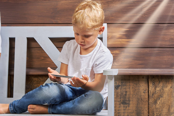Little boy in jeans and a white T-shirt sits on a wooden bench with a smartphone in his hands against the background of a wooden house. Concept of using gadgets by children - Zdjęcie, obraz