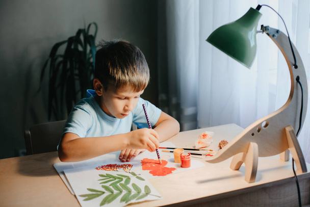 Söpö lapsi istuu pöydän ääressä ja piirtää rowanmarjoja albumilevylle kuivilla pihlajan lehdillä - Valokuva, kuva