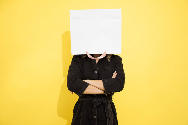 Photo of unrecognizable woman stands with shopping bag on head, stress relief with shopping, shopaholic, female psychology, studio portrait on yellow background.  - Foto, Imagen