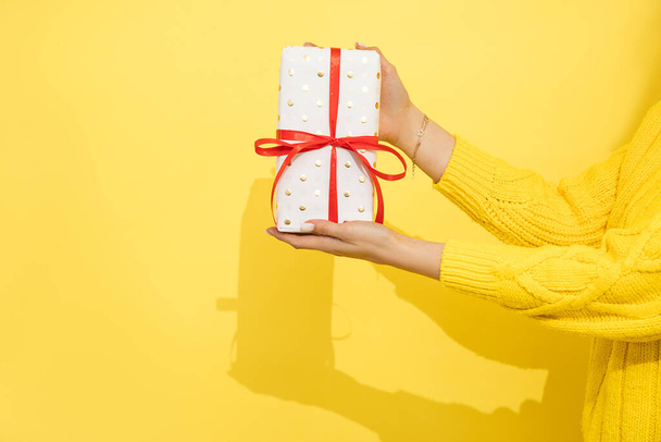 Unrecognizable Woman in sweeater holding in hand present, gift box with ribbon in wrapping paper on yellow background. Close-up photo, Gift Concept. - Photo, image