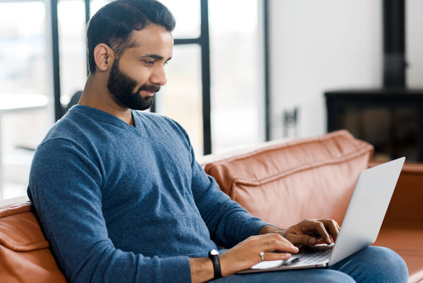 Handsome Indian guy in casual wear using a laptop sitting at home on the couch - Fotó, kép