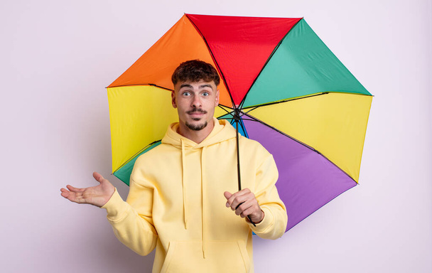 young handsome man feeling puzzled and confused and doubting. umbrella concept - Foto, Imagem