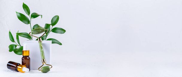 Jade crystal facial roller met bruine cosmetische flessen en groene bladeren, horizontaal, kopieerruimte - Foto, afbeelding