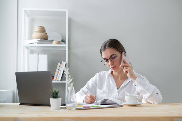 Business woman talking on phone and writing in notebook sitting her desk. - Photo, image