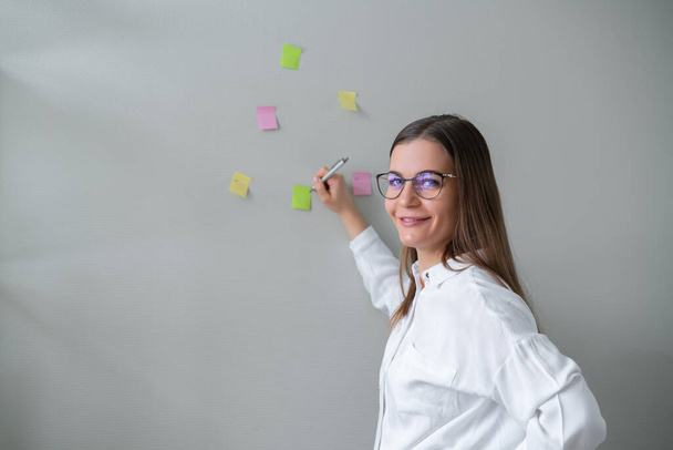 Young woman stands against wall and writes on colored stickers. Business planning. - Foto, Imagem