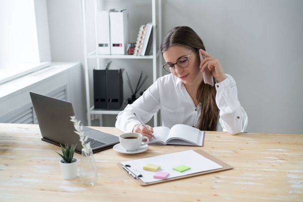 Young business woman talking on phone while sitting table  home, in front of  laptop. Writes information to work notebook. - Foto, Imagen