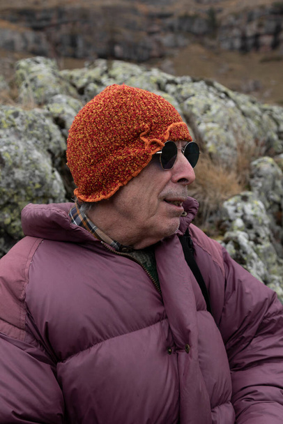 Um montanhista, homem adulto sênior, em seus 70 anos, em um casaco de penas, desfruta do cenário enquanto descansa apoiado em algumas rochas cheias de líquen, Pirinéus, Espanha - Foto, Imagem