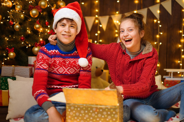Portrét chlapce a dívky v novoroční výzdobě. Otevřou krabici, zasmějí se a baví se. Dovolená světla, dárky a vánoční stromeček zdobený hračkami. - Fotografie, Obrázek