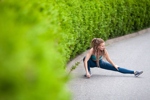 Sport girl with dreadlocks doing warm-up and stretching outdoors by the green fence. Sport and healthy lifestyle concept.  - Photo, Image