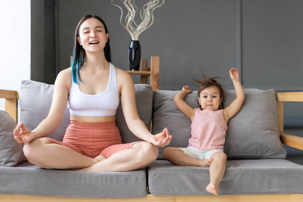 Young mom practices yoga on couch with her little daughter. Lotus position. - Фото, изображение