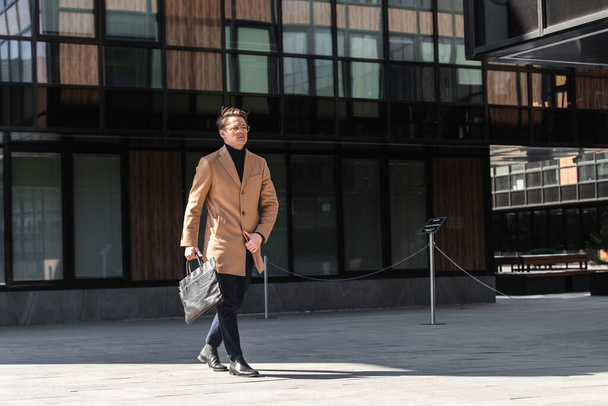 Young businessman in coat, glasses and with briefcase goes to a business meeting against the backdrop of modern business center. - Foto, immagini