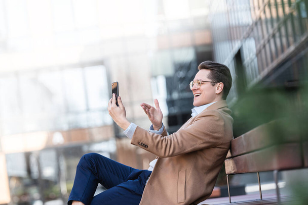 Young, confident, business man is talking on video call while sitting on street bench in courtyard of business center, gesturing to interlocutor, online conference. - Фото, зображення