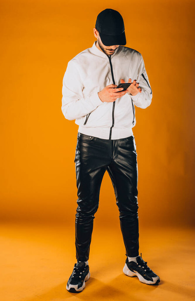 Cool young man dressed in white wind jacket posing in modern studio against orange background - Foto, imagen