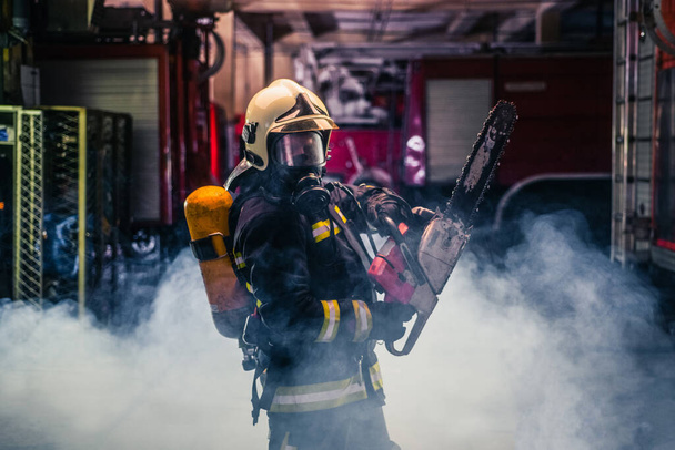 Portrait of young fireman standing and holding a chainsaw in the middle of the chainsaw's smoke - Photo, Image