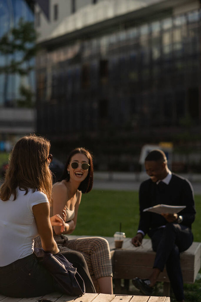 Τρεις πολυφυλετικοί επιχειρηματίες γελάνε με τα νέα σχέδια ενώ κάθονται στον πάγκο. - Φωτογραφία, εικόνα