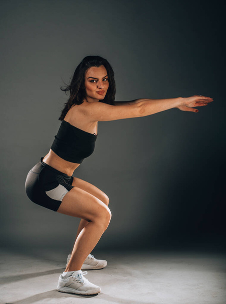 A dark-haired woman coach in a sporty short top and gym makes and stretch legs, doing exercise on a dark isolated background in studio - Zdjęcie, obraz