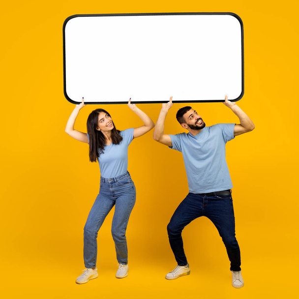 Cellphone display template. Arab spouses holding huge smartphone with blank copy screen over yellow background - 写真・画像