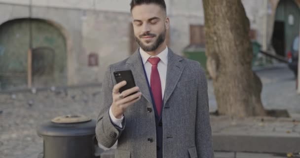 gelukkige jonge zakenman in pak houden mobiel, naar beneden kijken en glimlachen, knikken en poseren in Sibiu - Video
