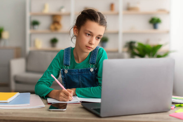 Dívka sedí za stolem, píše do učebnice pomocí PC - Fotografie, Obrázek