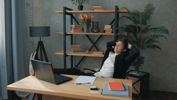 A young promising businessman drinks coffee carelessly with his legs on the table.Young attractive businessman is resting from work with his legs on the table. Young handsome businessman writes a financial report on a laptop and leans back in a chair - 写真・画像
