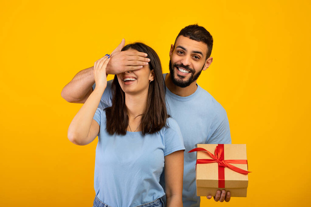 Boldog szülinapot vagy évfordulót. Boldog arab férfi takarja felesége szemét, így csomagolt ajándék doboz, sárga háttér - Fotó, kép