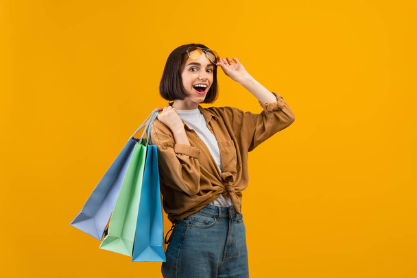 Speciální velký prodej na dovolenou. Radostná žena nosí sluneční brýle a drží barevné nákupní tašky, žluté pozadí - Fotografie, Obrázek