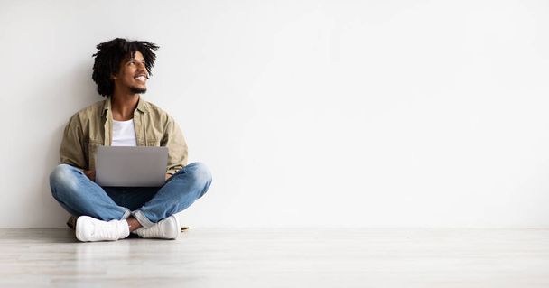 Mladý afroameričan sedí na podlaze s laptopem a dívá se jinam - Fotografie, Obrázek