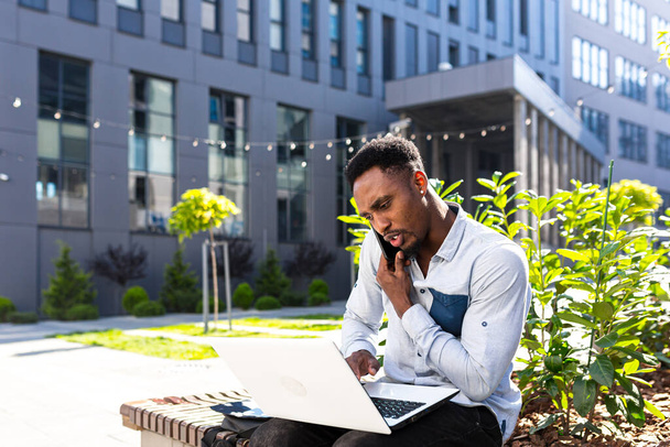 african american male freelancer working remotely online broker with exchanges stocks talking on mobile phone and laptop Merchant seller. Male in city park a bench modern urban background business man - Фото, изображение