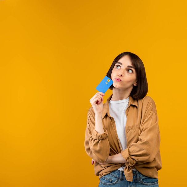 Unlimited shopping concept. Dreamy young woman holding credit card and thinking what to buy, yellow background - Photo, image