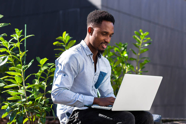 black man freelancer working online using laptop sitting on bench outside office modern building in city urban park on street. Happy African american guy student in casual outdoors typing on keyboard - Foto, immagini