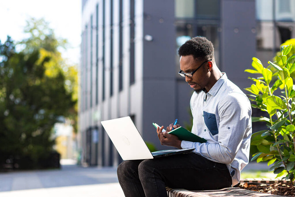 Young african american male student sitting in a city park on a bench with laptop and notebook studying online outdoors Man freelancer in glasses with books learn working remotely in street e-learning - Foto, Imagem