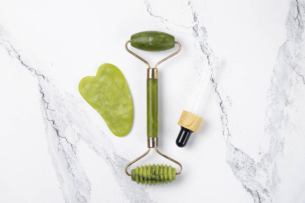 Set for home face care. Guasha and face massage roller made of green jade stone. Cosmetic lifting kit on a white marble background. - Zdjęcie, obraz
