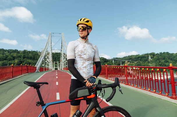 Portrait of a sporty man in a cycling outfit standing with a bicycle on the bridge and posing for the camera looking to the side. - Photo, Image