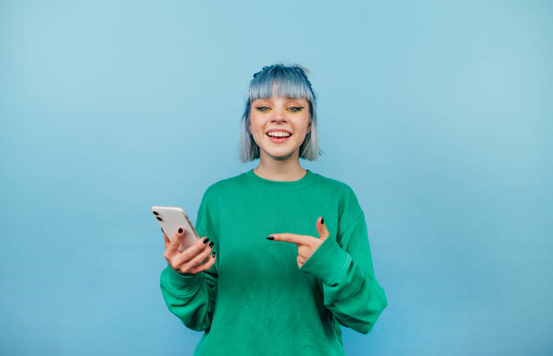 Positive helle Dame in grünem Sweatshirt und gefärbtem Haar steht auf blauem Grund, das Smartphone in der Hand und blickt mit einem Lächeln im Gesicht in die Kamera. - Foto, Bild