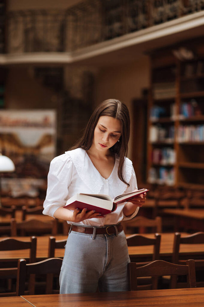 Une belle femme en chemisier se tient dans une bibliothèque atmosphérique publique et lit un livre avec un visage sérieux. - Photo, image