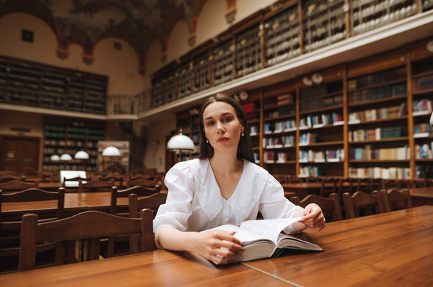Attraktive Frau sitzt mit einem Buch in der Hand in einer öffentlichen Bibliothek und posiert mit ernstem Gesicht für die Kamera - Foto, Bild