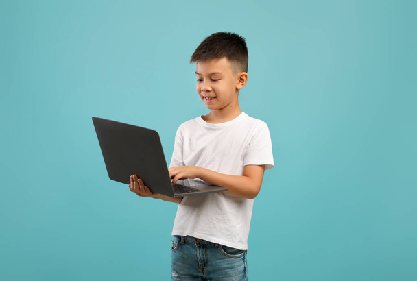Retrato de poco asiático chico usando portátil mientras de pie sobre fondo azul - Foto, Imagen