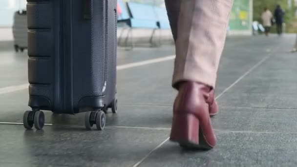 Vrouw in een lichte broek, kastanjebruine hielschoenen, met grijze koffer op wielen loopt langzaam naar buiten langs luchthaventerminal en lege banken, onderaanzicht van de benen. Reisconcept, zakenreis - Video