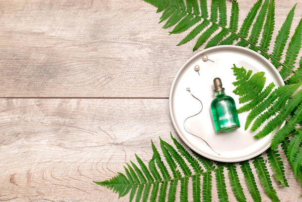 自然化粧品香水エッセンシャルオイルは、コピースペースと緑のシダの枝と緑のガラス瓶、プレート上の宝石アクセサリー、木製の背景に血清。生態系の美しさの概念. - 写真・画像