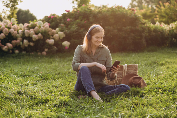 Retrato de una joven rubia feliz escuchando música con auriculares y sonriendo en una hierba verde al aire libre. Amante de la música disfrutando. Estilo de vida moderno. Concepto de educación en línea y remota - Foto, imagen