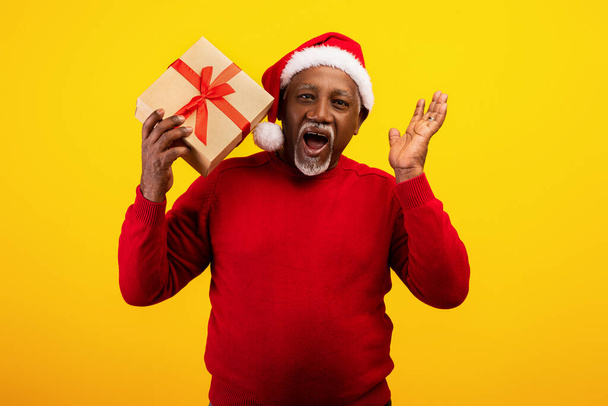 Podekscytowany starszy czarny mężczyzna w czerwonym swetrze i kapelusz Świętego Mikołaja trzymając pudełko z prezentami, krzycząc HO HO HO, oferując dużą wyprzedaż świąteczną - Zdjęcie, obraz