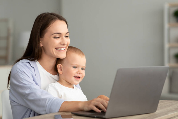 Portret van een gelukkige moeder met kleine baby op handen met behulp van laptop samen - Foto, afbeelding