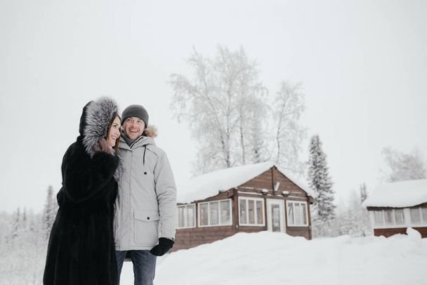 Όμορφο ερωτευμένο ζευγάρι αγκαλιάζει ο ένας τον άλλο, μικρό ξύλινο σπίτι και το χιόνι στην backgroun - Φωτογραφία, εικόνα