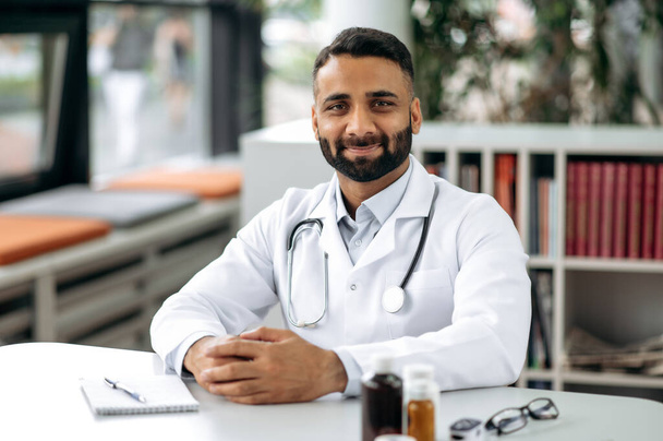 Portret przystojnego indyjskiego terapeuty generalnego, w mundurze medycznym i stetoskopie, siedzącego przy biurku w gabinecie, w szpitalu, patrzącego w kamerę, uśmiechniętego przyjaznego - Zdjęcie, obraz