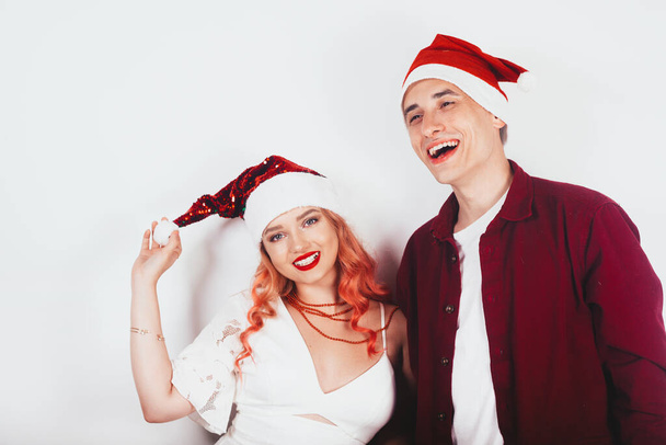 Couple in red santa hats on a white background laugh and celebrate new year and christmas. girl with pink hair and white dress laughs. - Photo, Image